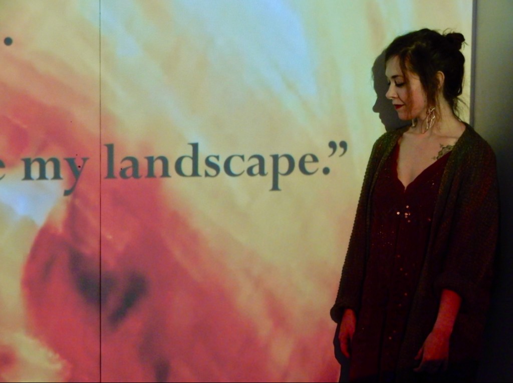 Maria, a white woman with her black hair in a bun, big earrings, and red lipstick, stands in front of letters on a screen that say "my landscape"- a video representation of her epileptic ElseSpace