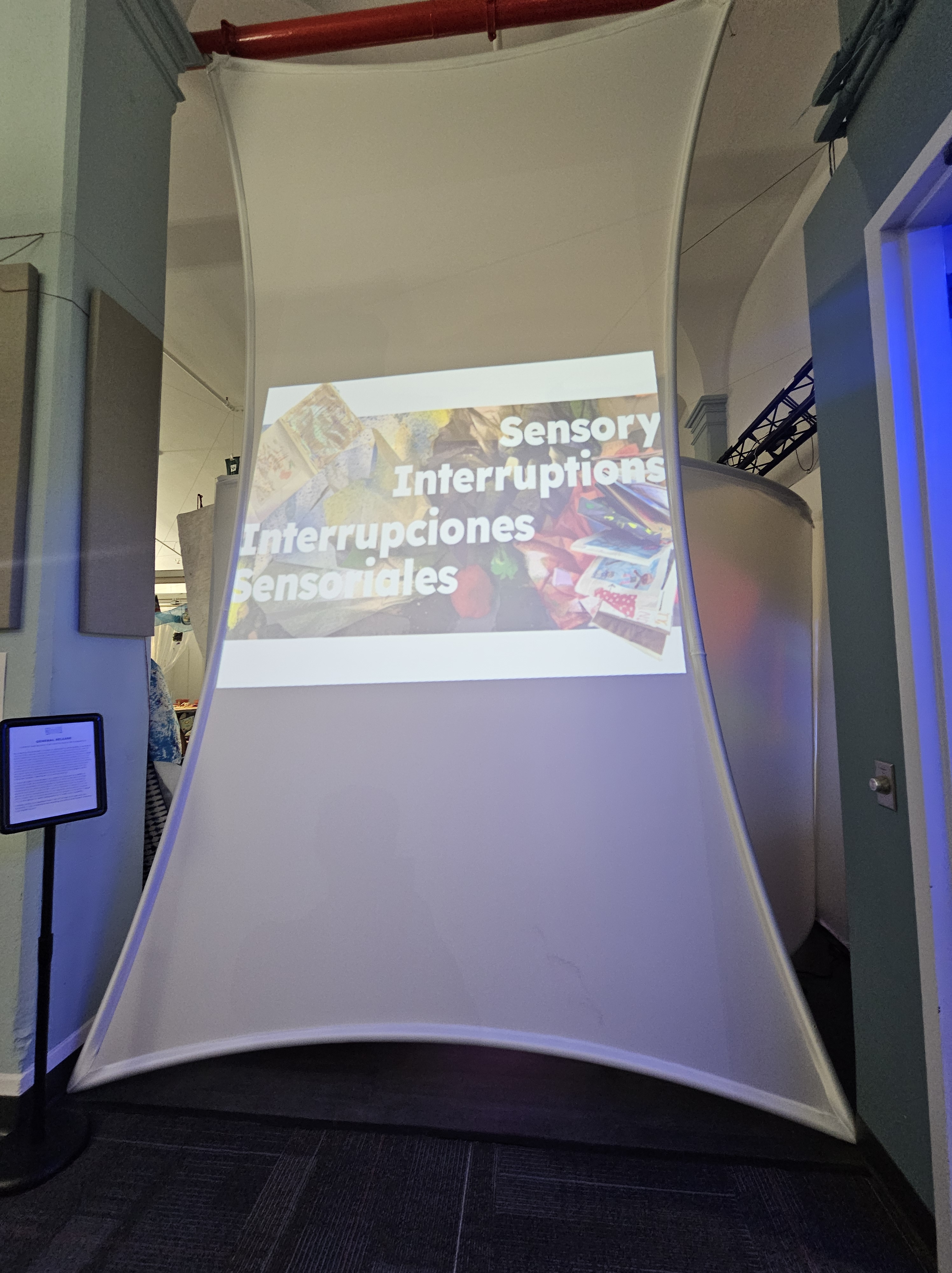 a photo of a large screen with the words SENSORY INTERRUPTIONS// INTERRUPCIONES SENSORIALES across unfolding artist books