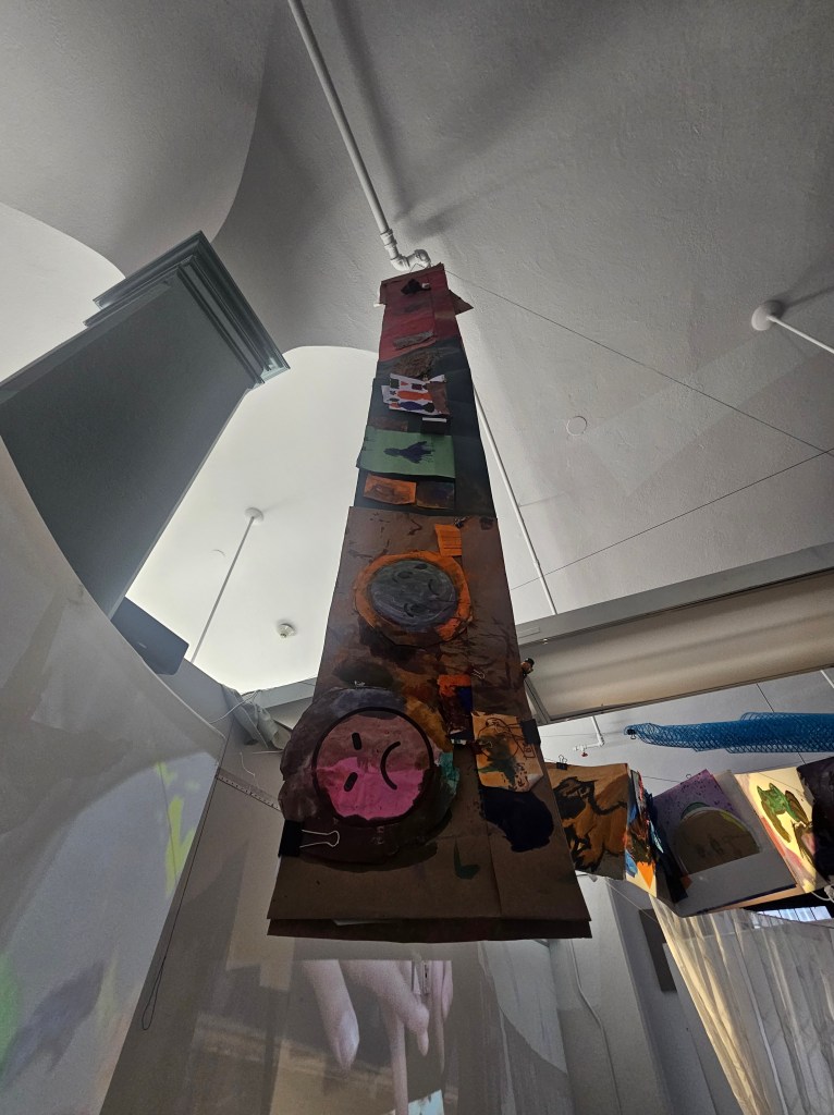 [a photo of another student accordion book hanging from the ceiling. Collaged images, paint, and pasted treasures can be seen peeking out.]