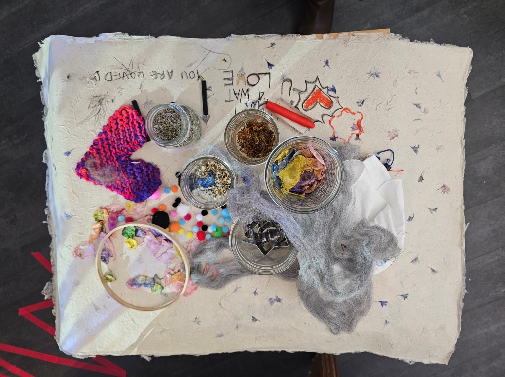 [a photo of participatory materials on a table of the exhibit. There are dried flower petals, lavender, seed pods, and fibers on top of handmade paper that is sketched and drawn on with crayon.]