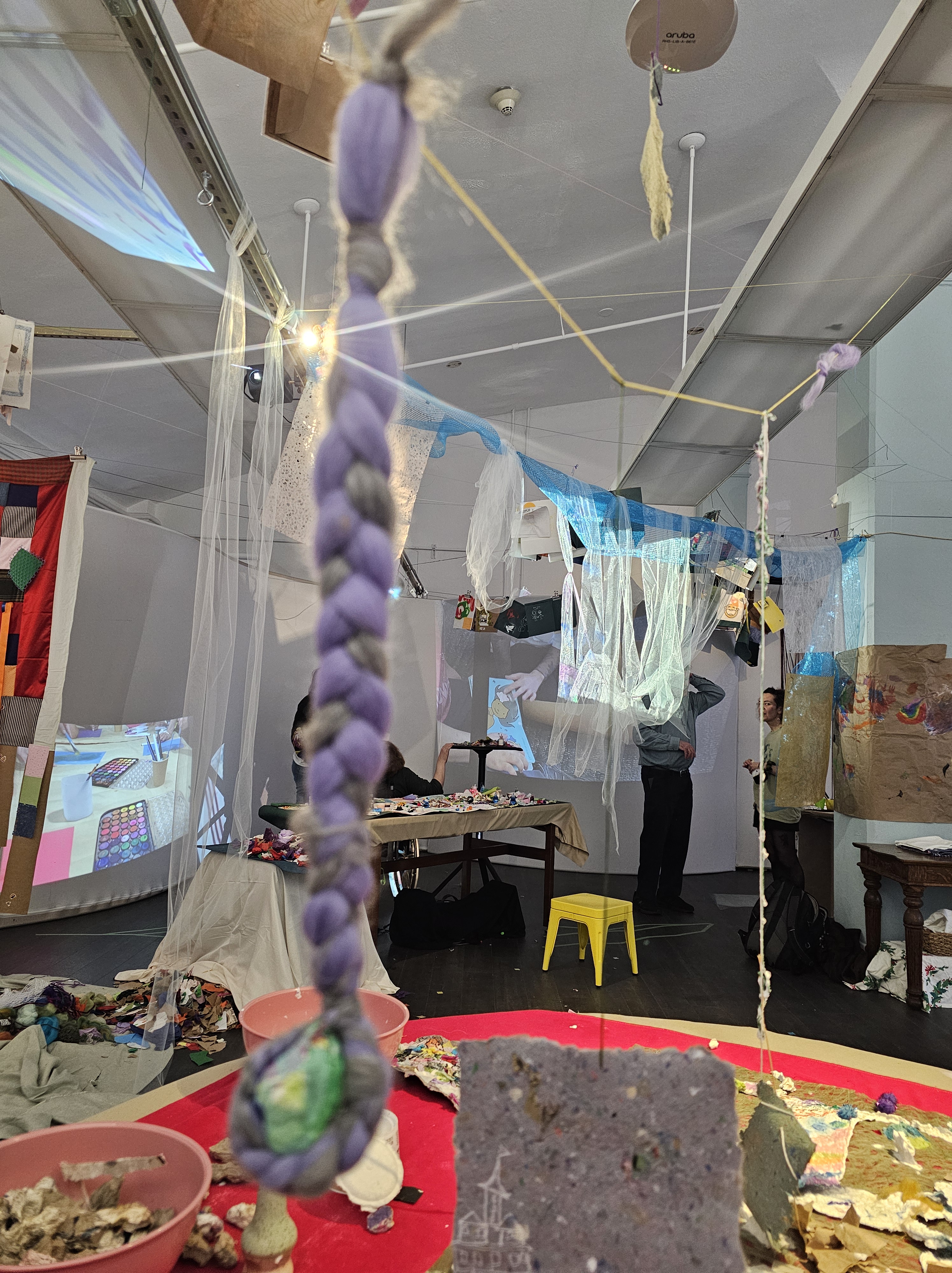 [a photo from the collaborative "overflow" paper pulp station. Pulp in various forms is scattered across the table, a braided piece of purple fiber hangs over, and there is light, sounds and energy moving through the space.]