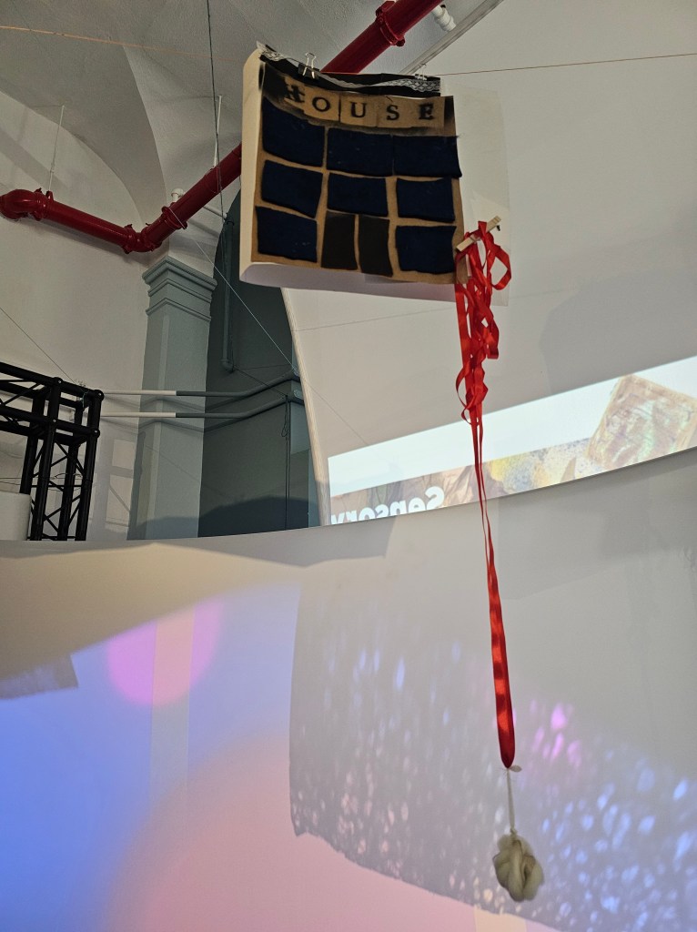 [a photo of a student artist book hanging from the ceiling. The cover contains cut out squares of fabric aligned in rows to create windows below the stenciled word "HOUSE". Behind the book light reflects and diffracts through windows of paper.]