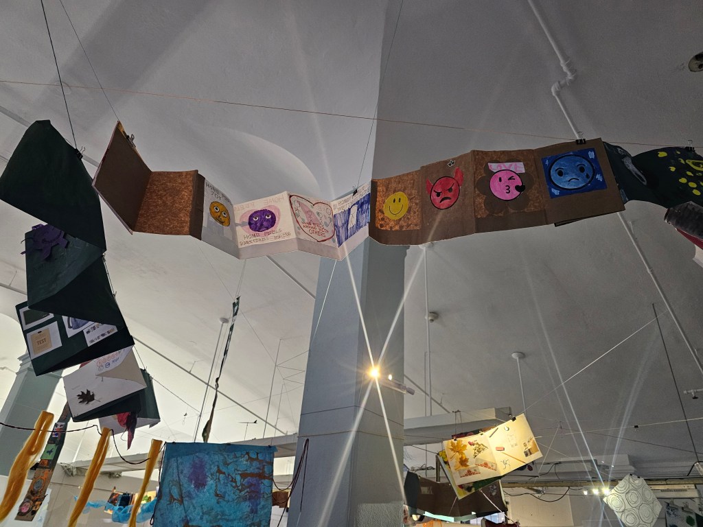 [A photo of student accordion books floating from the ceiling, their treasures and collages peeking out and hanging from their pages. Light pours through and fibers and papers hang down.]